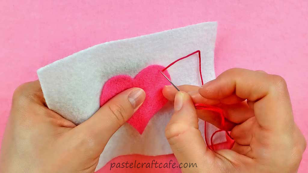 A needle about to sew the first stitch to attach a pink heart applique to white felt