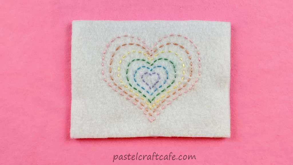 Six hearts in pastel rainbow colors is stitched onto a piece of white felt using the running stitch