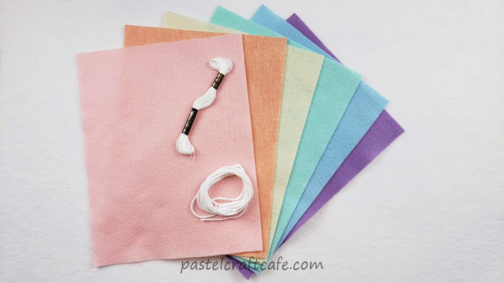 Six sheets of felt in pastel rainbow colors, a skein of white embroidery floss, and a small bundle of white cotton yarn