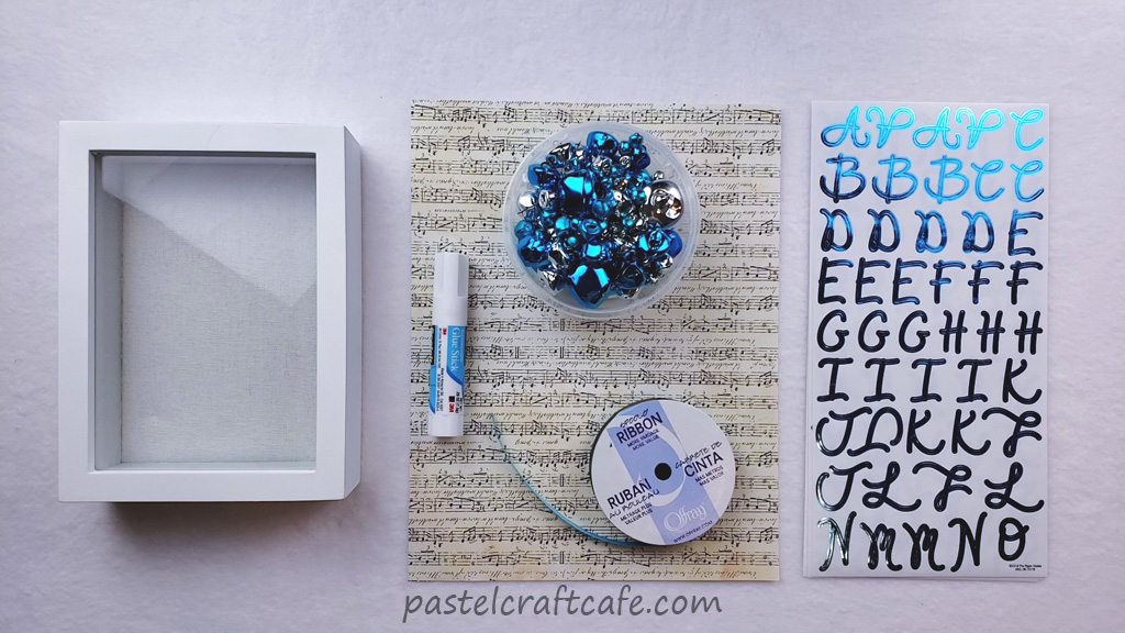 A white shadow box, scrapbook paper with a music sheet print, a glue stick, assorted size jingle bells in blue and silver, a spool of turquoise ribbon, and blue metallic alphabet stickers