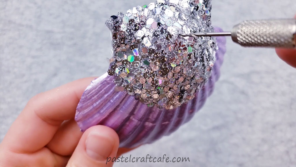 A hand drill clearing the drill hole on a glittered purple seashell ornament