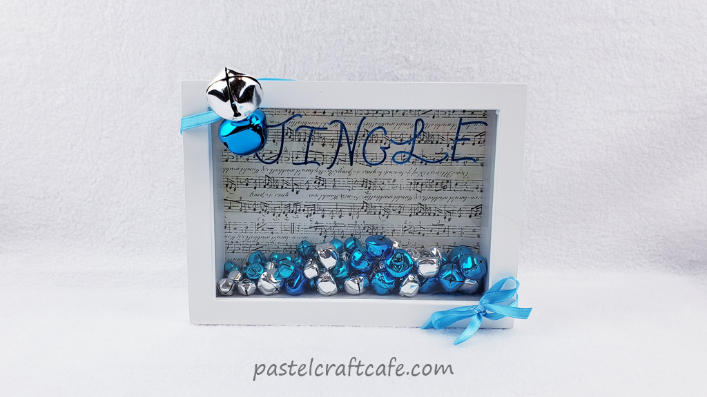 White framed shadow box filled with jingle bells and sheet music scrapbook paper with "JINGLE" on glass and bells and ribbon wrapped around frame