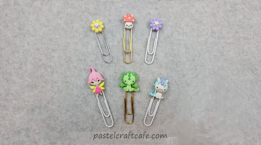 A set of six button paperclip bookmarks with various fantasy characters attached: a smiling yellow flower, a smiling mushroom, a smiling purple flower, a fairy, a dragon, and a unicorn
