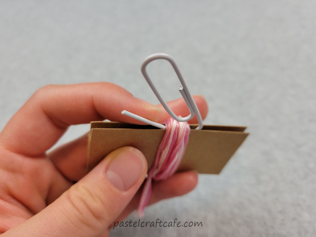 The open end of a paperclip being threaded onto a bundle of embroidery floss on a DIY mini tassel maker