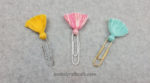 Three tassel paperclip bookmarks. A yellow tassel on a silver paperclip, a multicolored pink tassel on a white paperclip, and a blue tassel on a gold paperclip