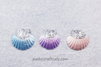 Three seashell ornaments with glitter on top in the colors of metallic blue, purple, and champagne pink