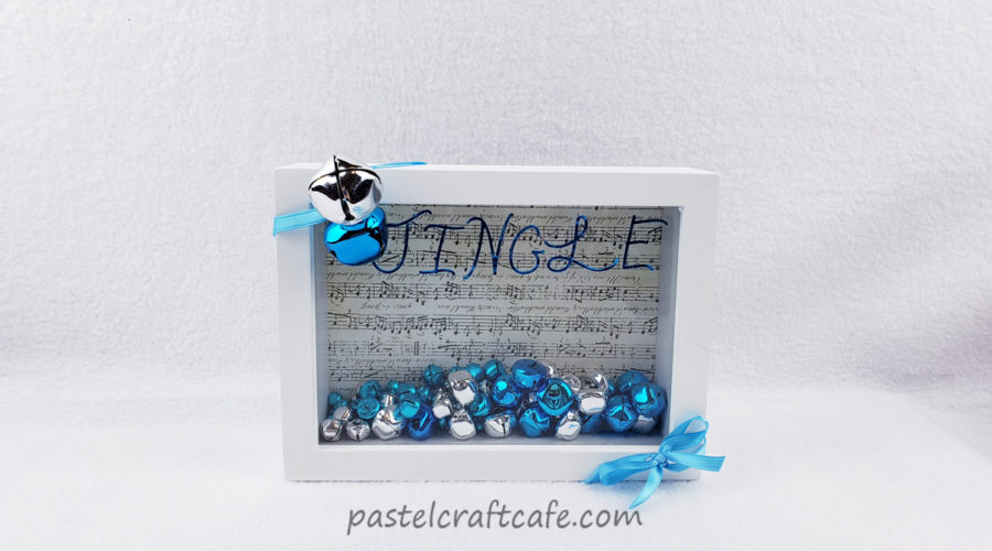 A shadowbox with a white frame wrapped in a ribbon with jingle bells and a bow attached. On the top of the glass are blue stickers that write out "Jingle" and inside the box are blue and silver jingle bells with sheet music backing