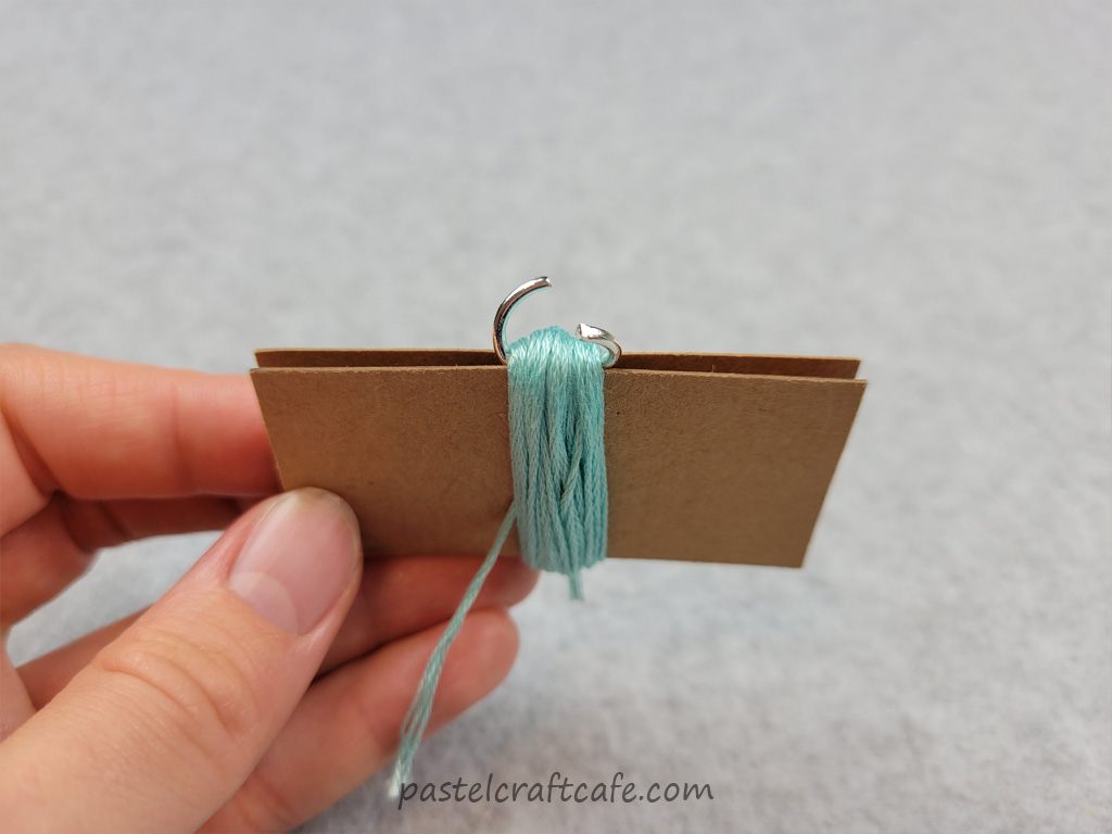 An open jump ring threaded under an embroidery floss bundle that is wrapped around a DIY mini tassel maker