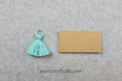 a finished tassel charm made of blue embroidery floss next to a rectangular DIY tassel maker
