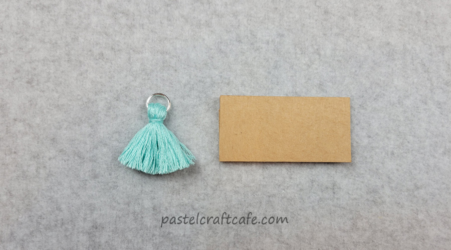 a finished tassel charm made of blue embroidery floss next to a rectangular DIY tassel maker