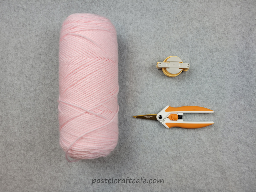 a skein of pink yarn, a pom pom maker, and scissors