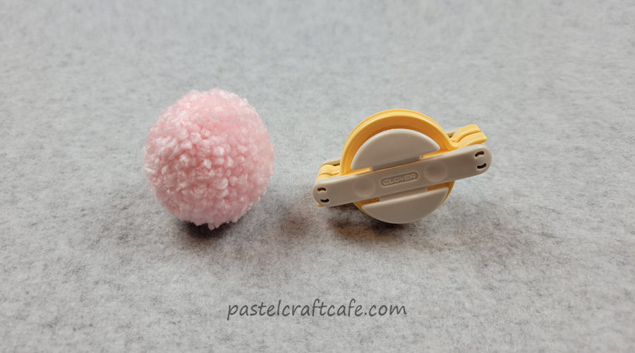 a small pink pom pom next to a yellow plastic Clover pom pom maker