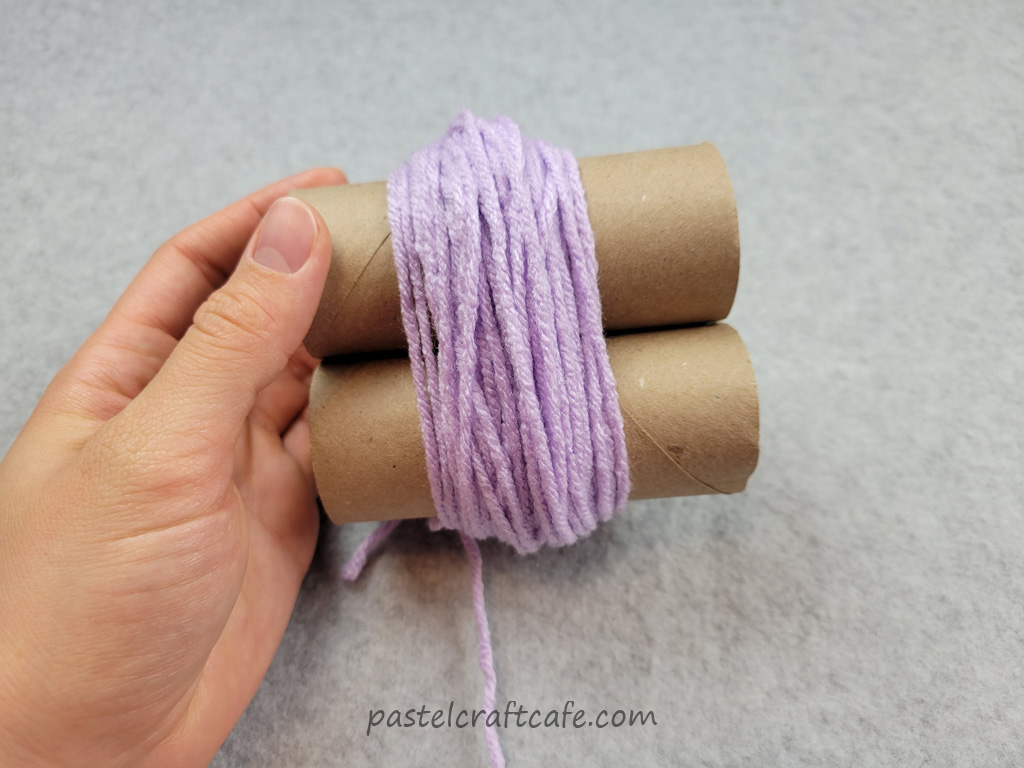 A hand holding two empty toilet paper tubes with a small bundle of yarn wrapped around them