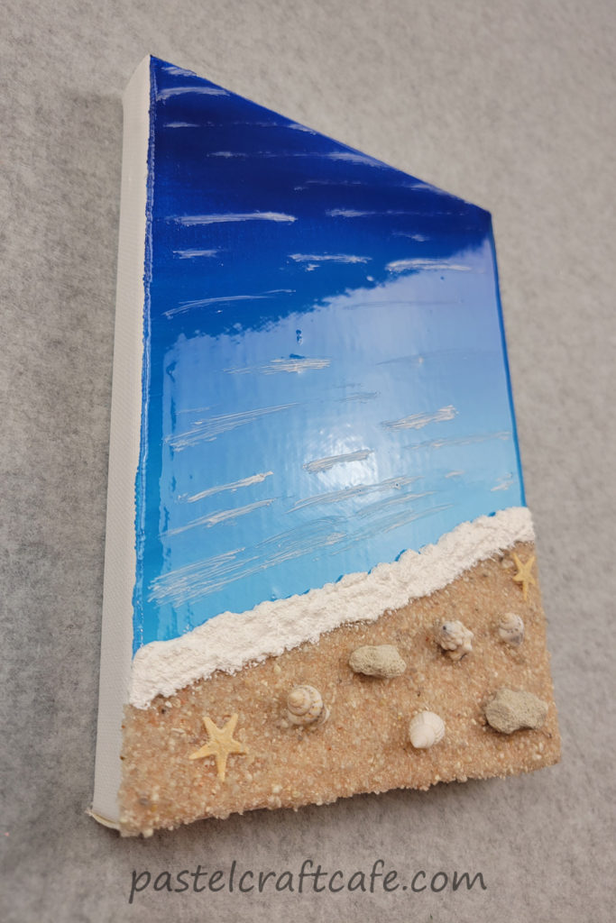 A mixed media beach canvas painting seen from an angle to show off the gloss on the water