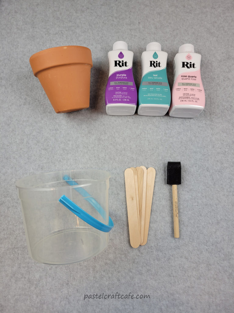 A terra cotta pot, three bottles of Rit dye, a plastic bucket, popsicle sticks, and a foam brush