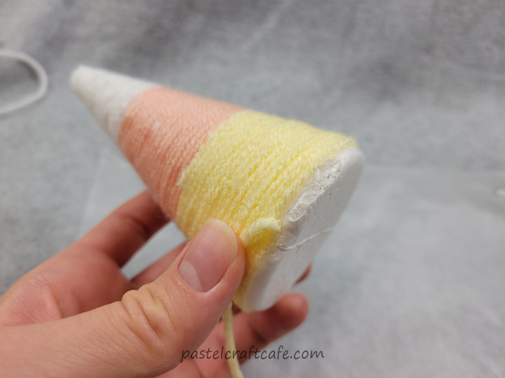 A styrofoam cone wrapped with white, orange, and yellow yarn in sections and the last line of yellow yarn being attached with hot glue at the bottom