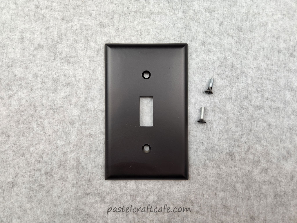 A lightswitch plate and two small screws that have been spray painted with black matte paint