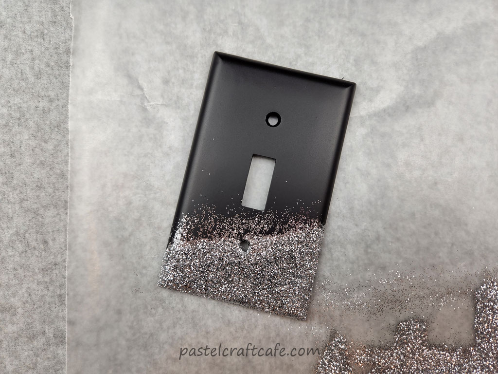 A black lightswitch plate that has the bottom edge decorated with a messy layer of fine silver glitter