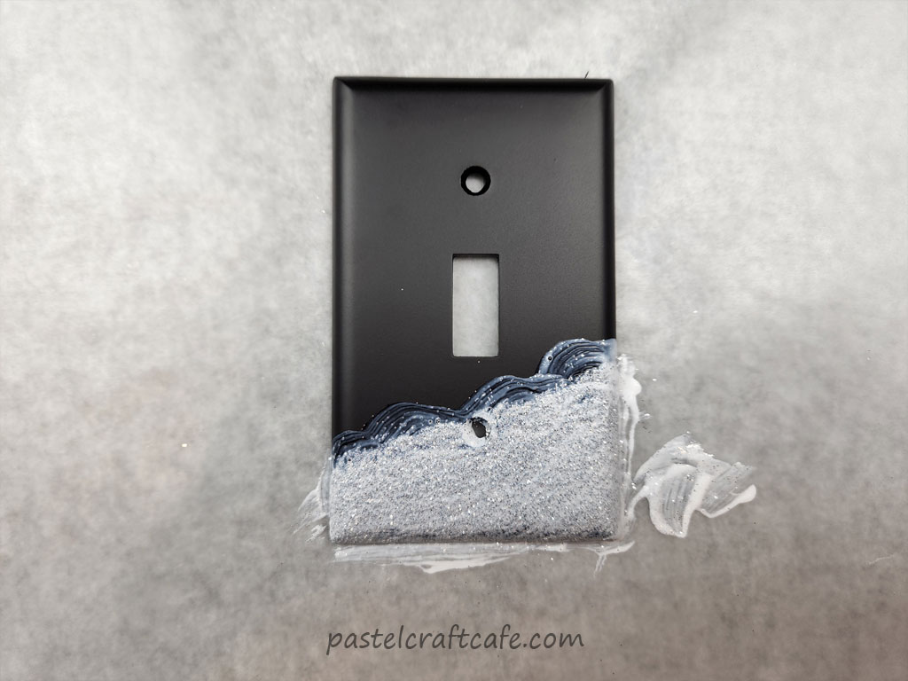 A layer of Mod Podge painted over the silver glitter that is decorating the bottom end of a black lightswitch plate