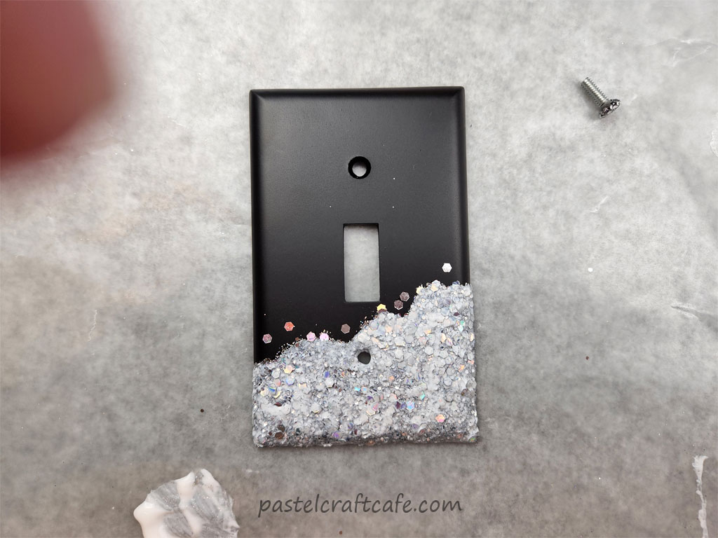 A layer of glossy Mod Podge painted over the silver holographic glitter that is decorating the bottom end of a black lightswitch plate