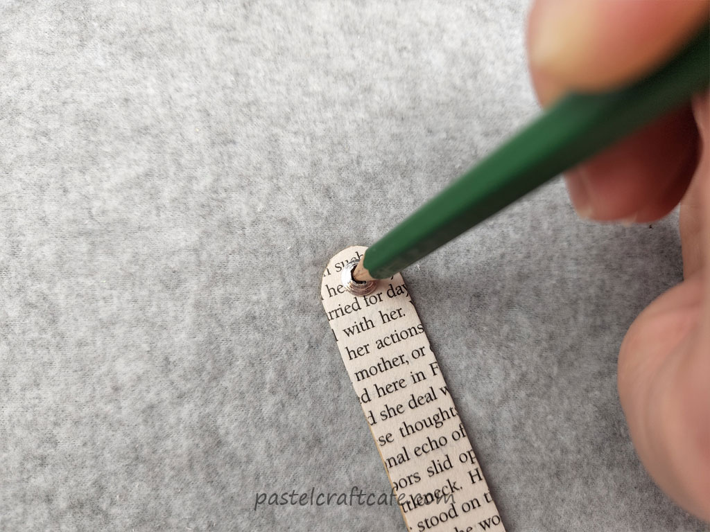 A pencil making a small mark at the top of a popsicle stick covered in book pages