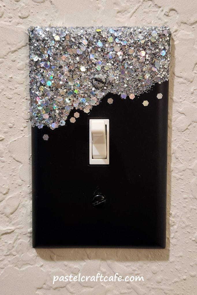 A light switch plate with silver holographic glitter decorating the top and black spray paint on bottom