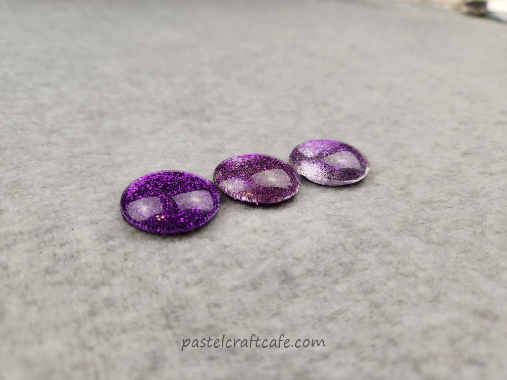 Three glass cabochons decorated with glitter on the backs. The adhesives used were Mod Podge, Gloss Varnish, and Clear Nail Polish. The three cabochons are seen at an angle to show Mod Podge dries the most clear and nail polish the least