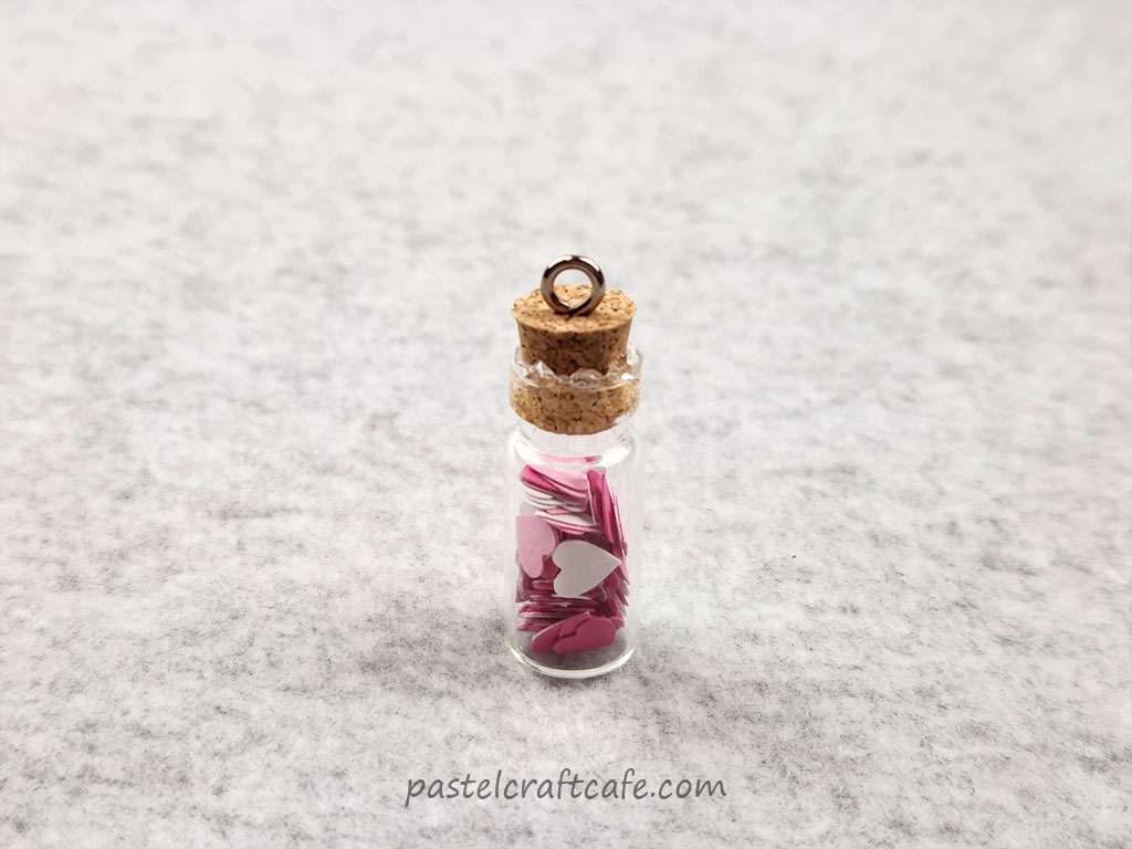 A tiny glass bottle charm with hearts in it. Excess glue is on the bottle where it meets the cork